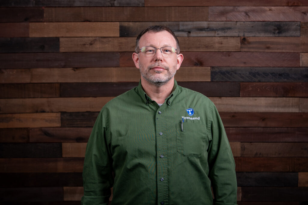 older man with glasses and a beard wearing a green shirt in front of a wooden background