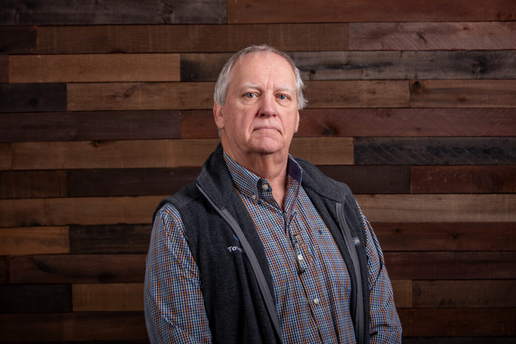 older man with a blue and orange button up shirt and a black vest in front of a wooden background