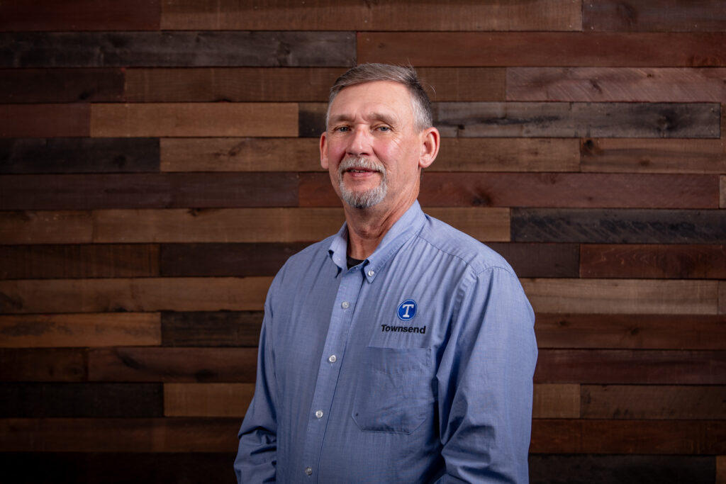 older man with a beard wearing a light blue shirt in front of a wooden background