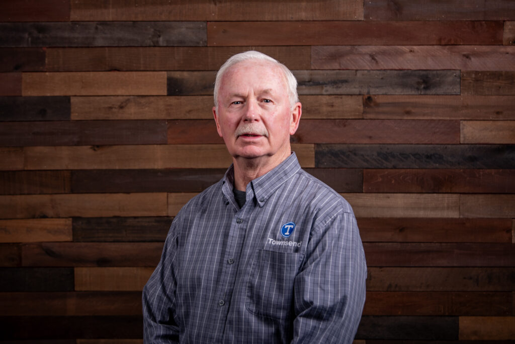 older man with a moustache and a blue button up shirt in front of a wooden background
