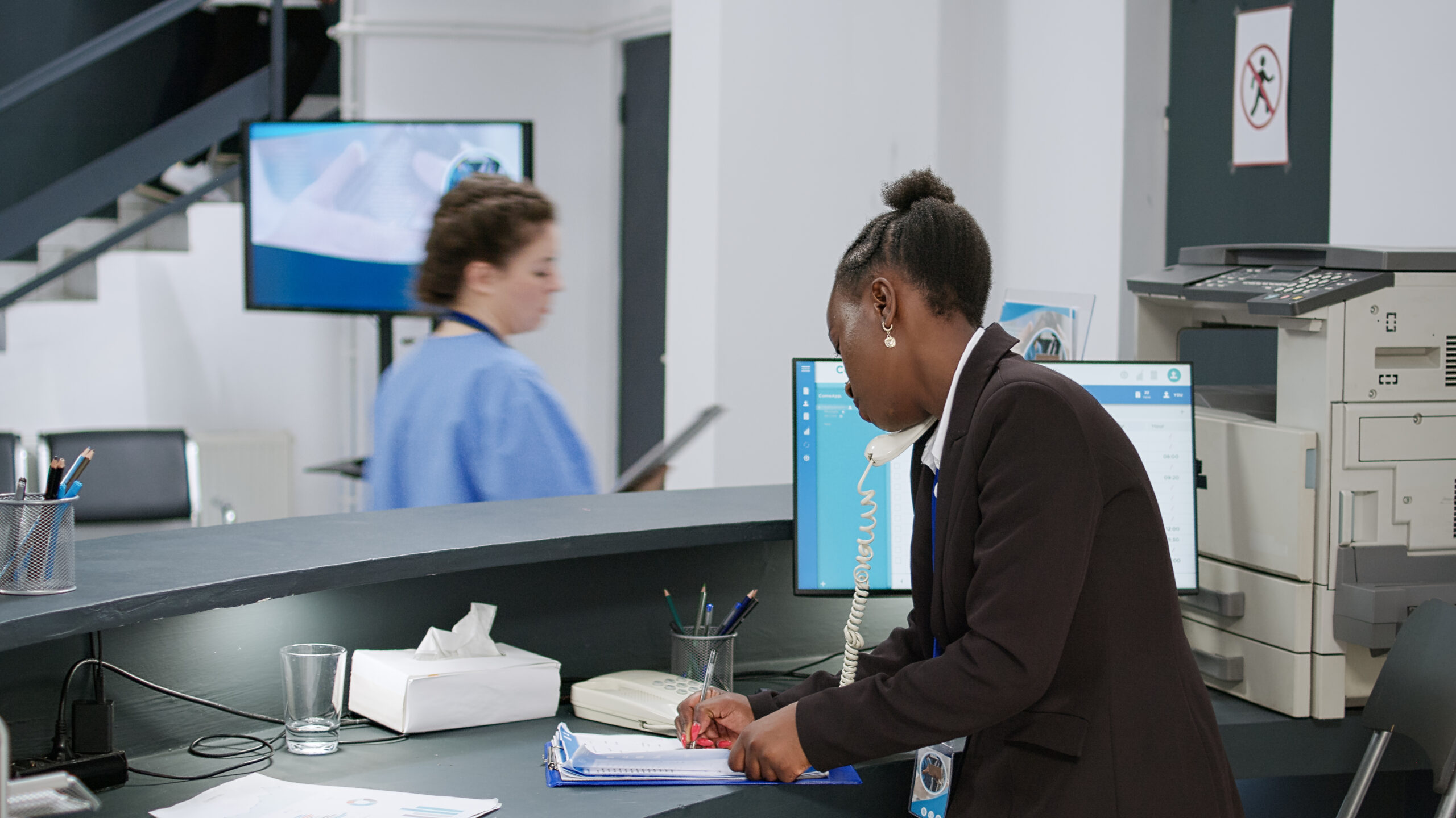 Black woman receptionist answering a landline telephone in a hospital with a dark hair, white medical professional passing by in the background