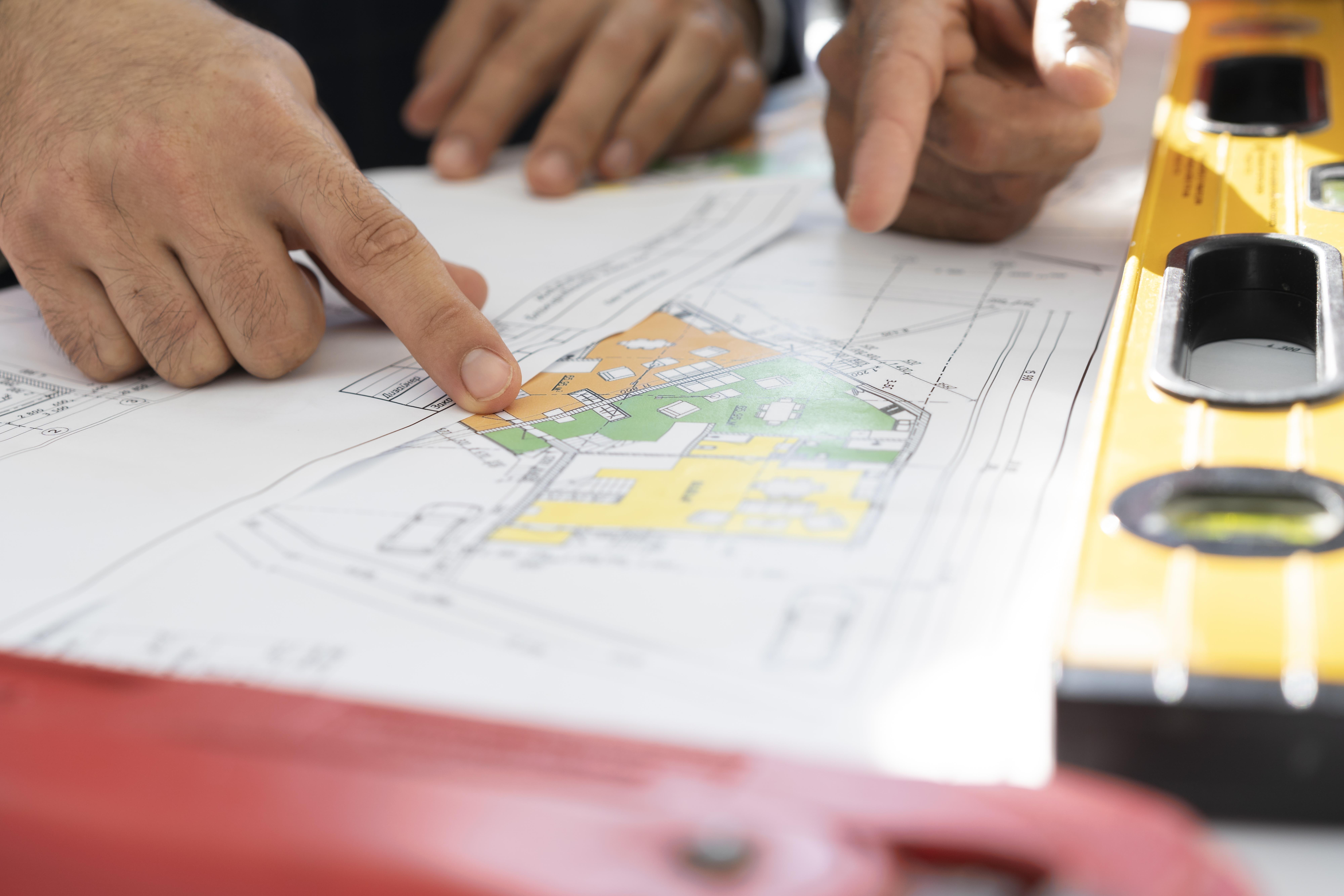 close up of paper building plans with several hands pointing at them and measurement tools on a table