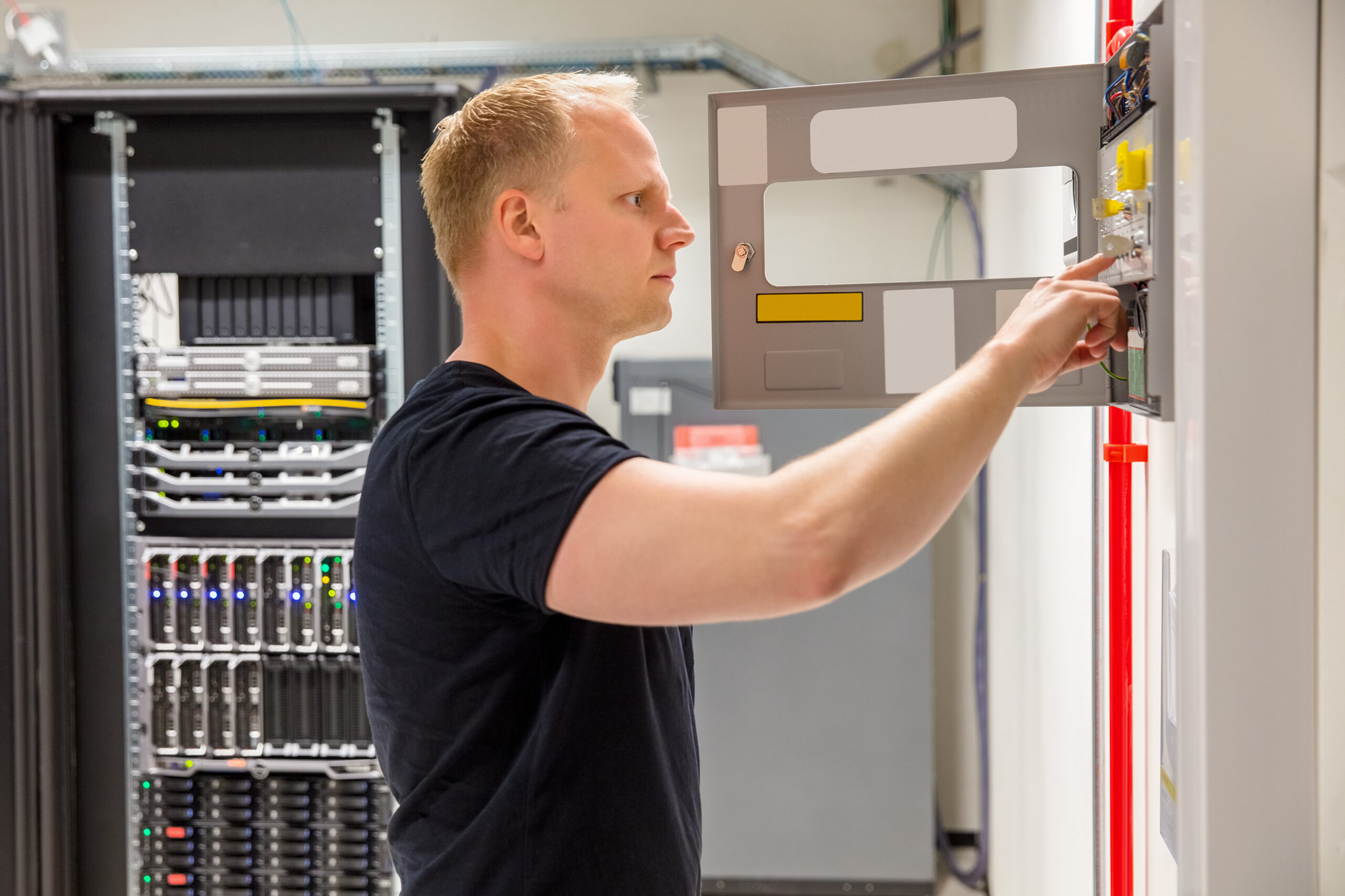 man in black shirt fixing an electrical unit on a grey wall with a bigger electrical unit behind him in the background