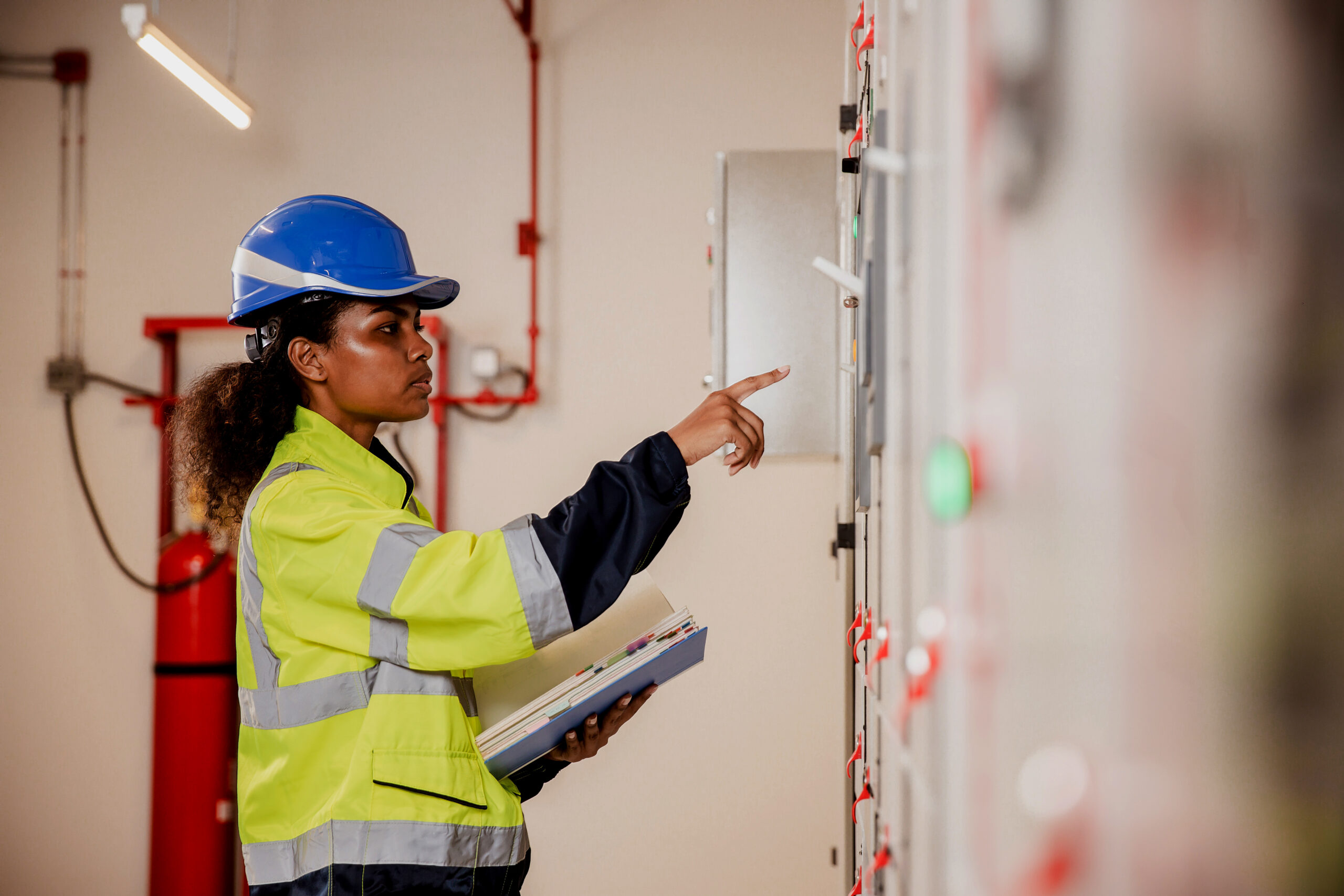 Black female electrical engineer wearing a yellow hi-vis jacket and a blue hardhat analyzing controls on a control panel