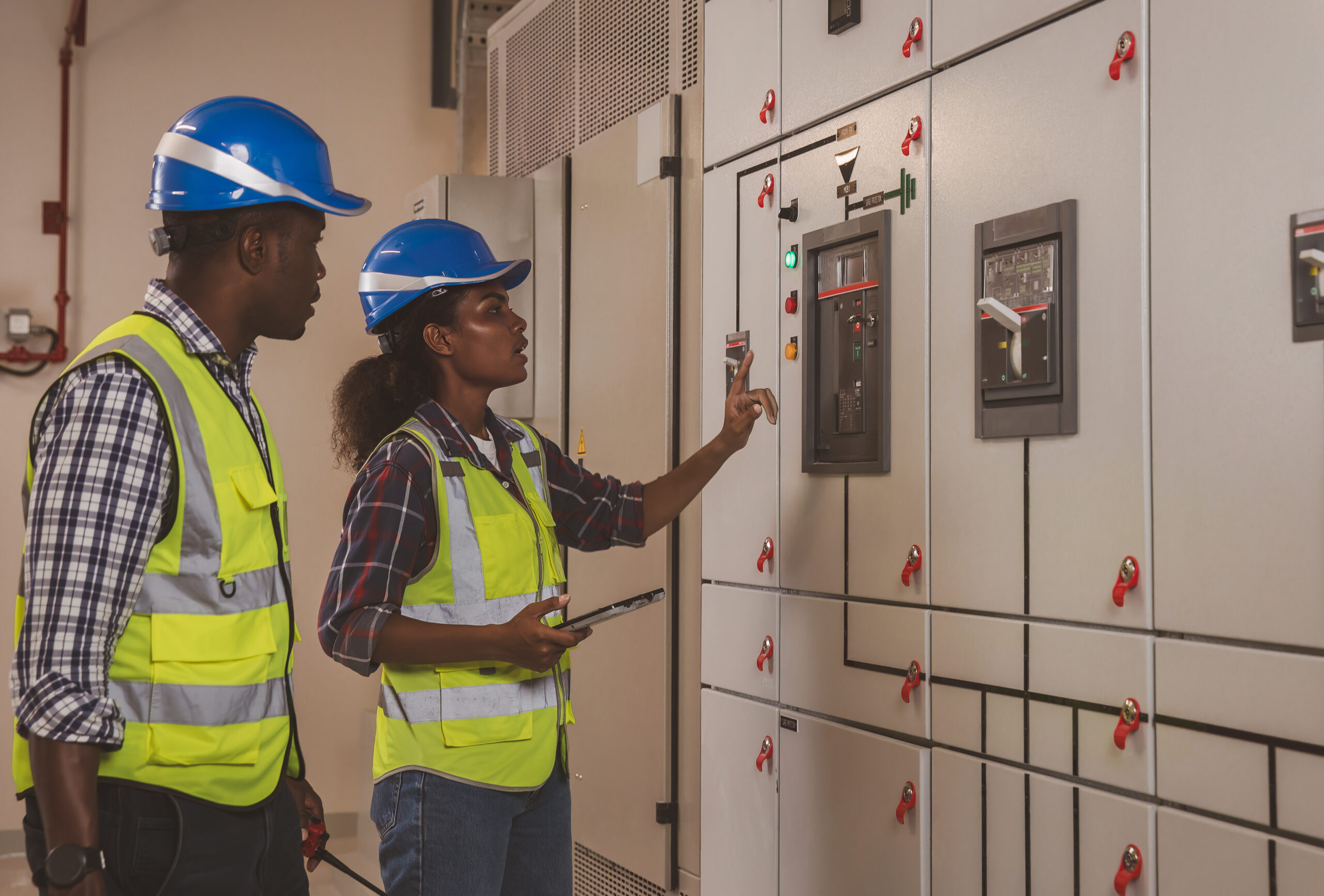 Two black electrical engineers working in a control room on a grey panel with multi colored buttons
