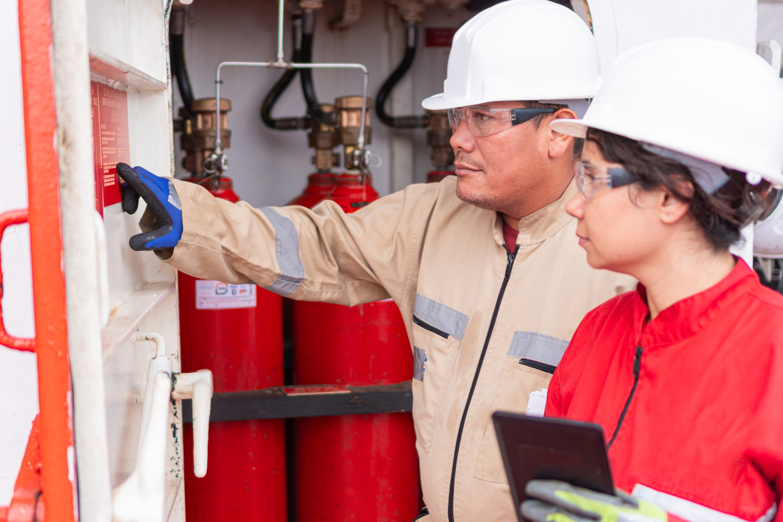 Male industrial professional wearing khaki jumpsuit, safety glasses, and white hard hat and female industrial professional wearing red jumpsuit, safety glasses, and white hardhat gauging metrics on the wall on site with red tanks behind them