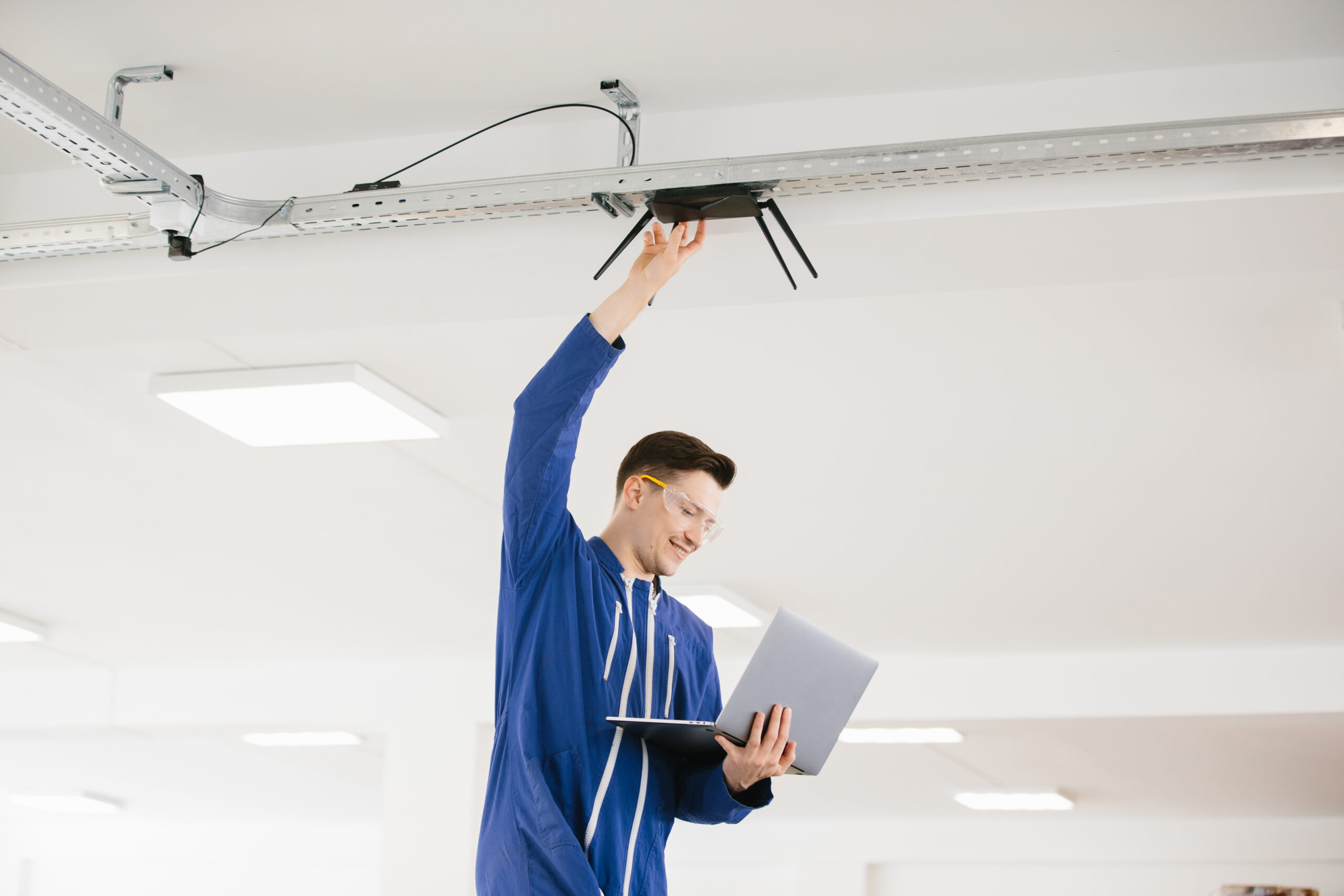 Young technician wearing a blue jacket and safety glasses installing a wireless router on a metal ceiling track