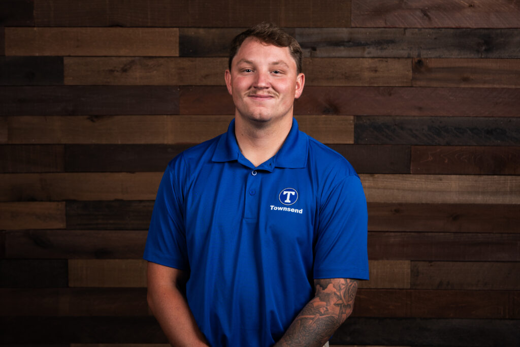 man with a moustache and sleeve tattoo wearing a blue polo in front of a wooden background