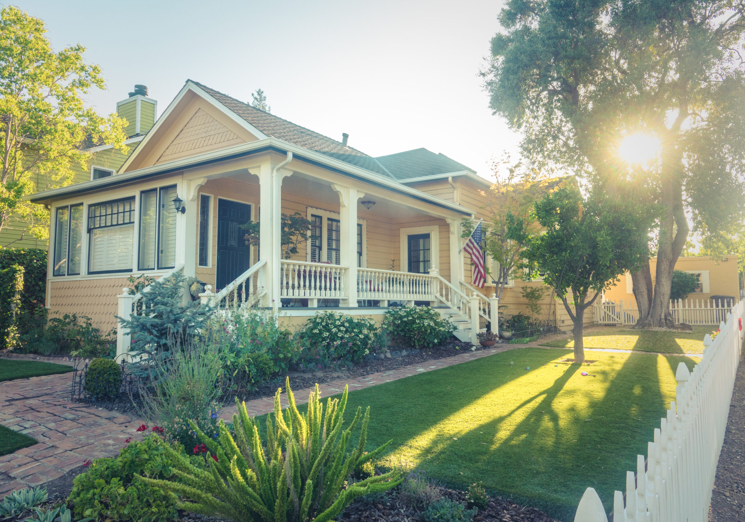 Traditional American suburban home painted yellow with white trim, an American flag, lush green landscaping, a white picket fence, and bright sunshine