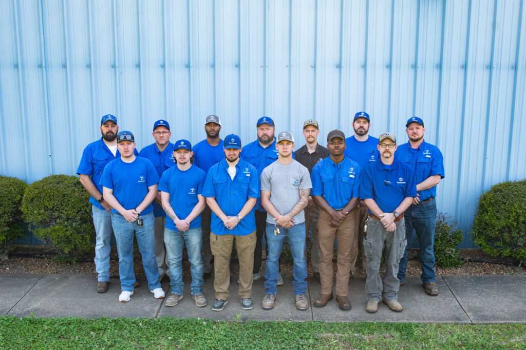 Group photo of Townsend systems team, most in blue, short sleeve work shirts and ball caps, in front of a metal wall, standing on a sidewalk