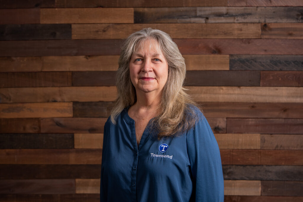 Middle age woman with long brown grey hair wearing a long sleeved blue Townsend Systems shirt in front of a wooden wall