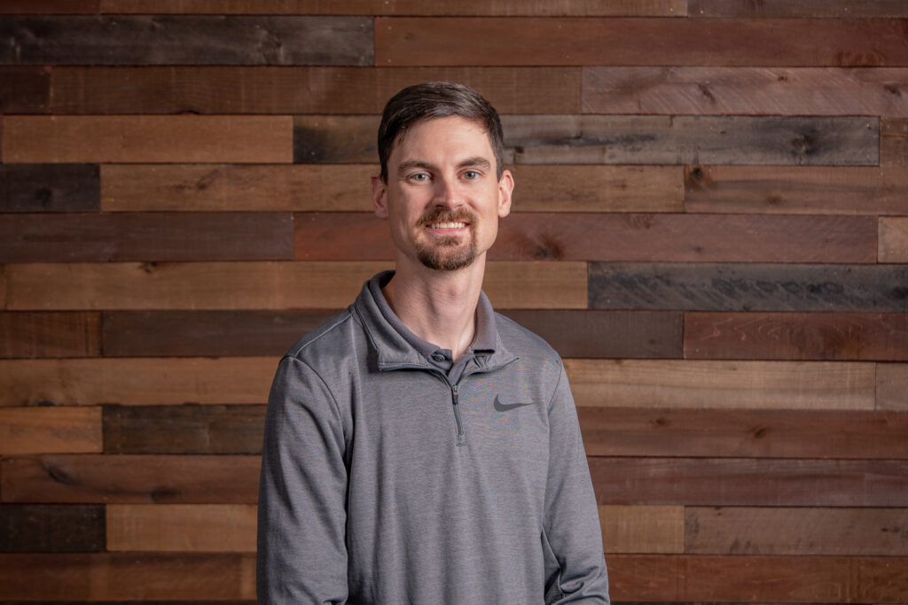 Young man with short brown hair and short facial hair wearing a grey Nike long sleeve shirt in front of a wooden wall